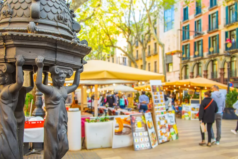 Close-up of decorative water fountain on La Rambla pedestrian street in Barcelona, Spain with blurred pedestrians in background.